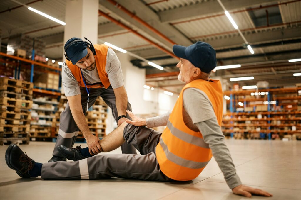 Young worker assisting his colleague with leg injury while working at distribution warehouse.